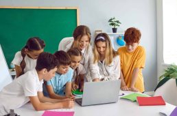 Smart children pupil boys and girls stand in front of laptop with woman teacher and view media material or online presentation during lesson in primary school classroom. Curiosity, education