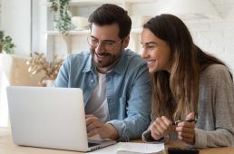 Happy young couple husband and wife using laptop computer looking at screen pay bills online in app calculate mortgage investment payment on website planning budget discuss finances sit at home table